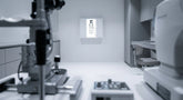 A professional ophthalmic exam room with an illuminated ETDRS visual acuity chart cabinet mounted on the wall under precise clinical lighting.
