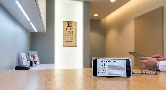 A sharply focused near vision testing card in the foreground of a modern optometry exam lane, with a blurred distance eye chart visible in the background.